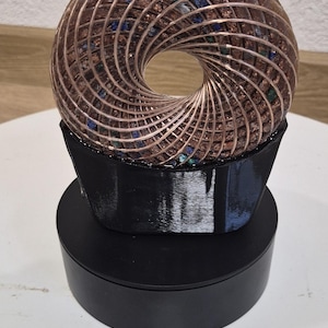 Pode incluir: Uma escultura em forma de toroide de cor cobre com um buraco central, repousando sobre uma base preta em camadas. O interior da escultura é preenchido com pequenas pedras coloridas. A obra de arte é exibida sobre uma superfície branca.