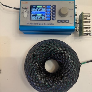 Pode incluir: Um gerador de sinal de 2 canais azul e preto com um logotipo de fantasma assustador. O dispositivo tem um botão para ajustar a frequência e botões para configurar a saída. O dispositivo está conectado a um objeto circular preto e verde com um furo no centro.