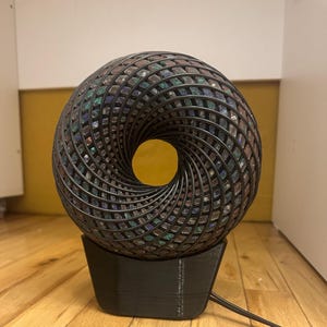 Könnte beinhalten: Dekorative Lampe mit Spiraldesign. Die Lampe hat einen dunklen, geometrischen Rahmen, der mit schillernden, mehrfarbigen Steinen gefüllt ist. Sie steht auf einem schwarzen Sockel und hat ein Netzkabel.