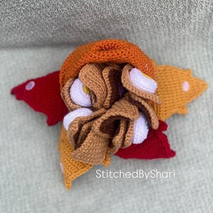 Pode incluir: Um taco de croch&ecirc; feito &agrave; m&atilde;o com elementos marrons, laranja e brancos, repousando sobre uma base em forma de estrela vermelha e amarela. O taco apresenta uma casca marrom, recheios brancos e amarelos e um topo laranja. O texto "StitchedByShari" &eacute; vis&iacute;vel.