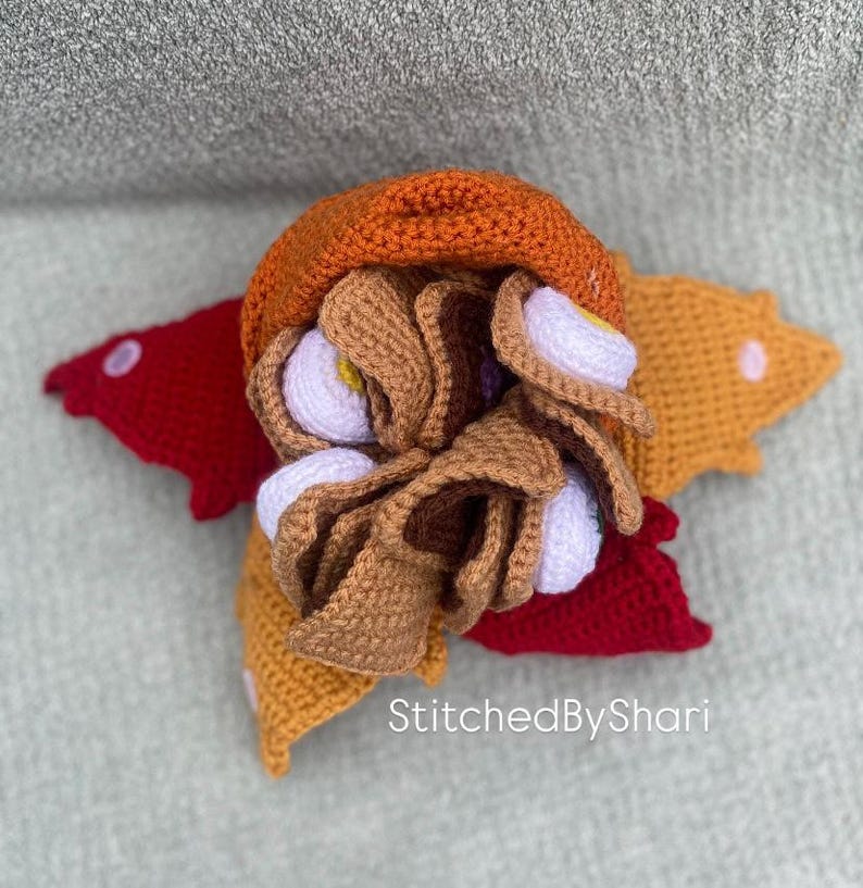 Pode incluir: Um taco de croch&ecirc; feito &agrave; m&atilde;o com elementos marrons, laranja e brancos, repousando sobre uma base em forma de estrela vermelha e amarela. O taco apresenta uma casca marrom, recheios brancos e amarelos e um topo laranja. O texto "StitchedByShari" &eacute; vis&iacute;vel.
