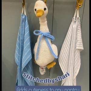 May include: A handmade crochet goose with a yellow beak and feet, and a blue bow, hangs from a brass rod between two blue and white striped towels. The text "Stitchedbyshari" and "Adds cuteness to any pantry" are visible.