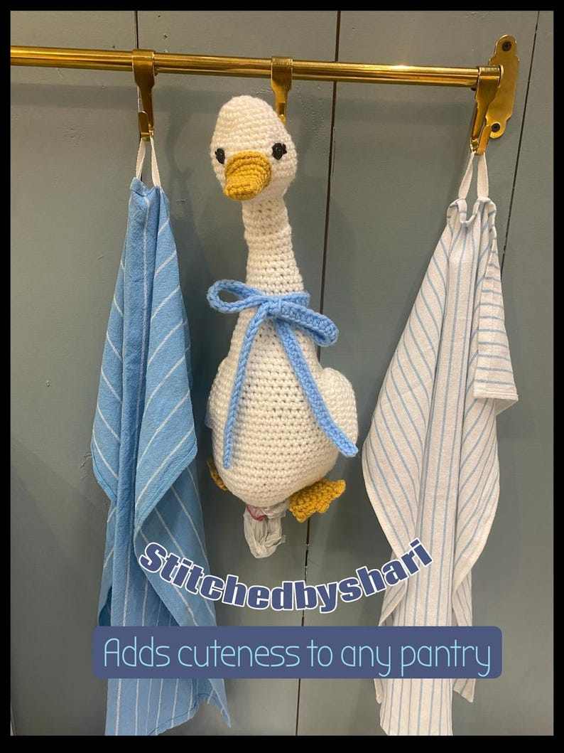 May include: A handmade crochet goose with a yellow beak and feet, and a blue bow, hangs from a brass rod between two blue and white striped towels. The text "Stitchedbyshari" and "Adds cuteness to any pantry" are visible.