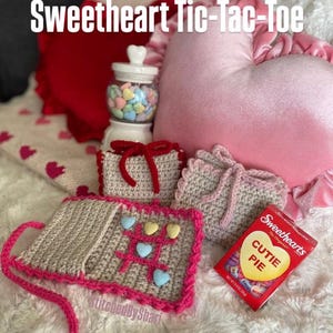 May include: A crochet tic-tac-toe game with a pink trim and heart-shaped pieces, alongside a box of Sweethearts candies and heart-shaped cushions. The image features the text "Sweetheart Tic-Tac-Toe" at the top, and "Stitched By Shari" at the bottom.