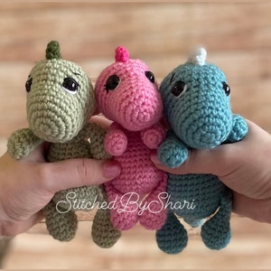 May include: Three crocheted dinosaur toys in green, pink, and blue. The toys have black eyes and are holding hands.