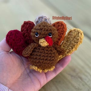 May include: A handmade crochet turkey in shades of brown, red, orange, and yellow. The turkey has a red wattle, a yellow beak, and black eyes. The text "StitchedByShari" is visible in the upper right corner.