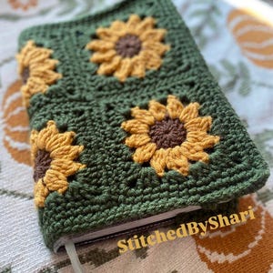 May include: Handmade crochet book cover in olive green with three sunflower appliques. The sunflowers have yellow petals and brown centers. The cover has a textured appearance and a light-colored ribbon bookmark. The text "StitchedByShari" is visible.