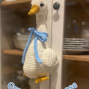 May include: A handmade, crocheted grocery bag goose in white with a yellow beak and feet. It has a light blue bow around its neck. The text "StitchedByShari" and "Grocery Bag Goose" are visible.
