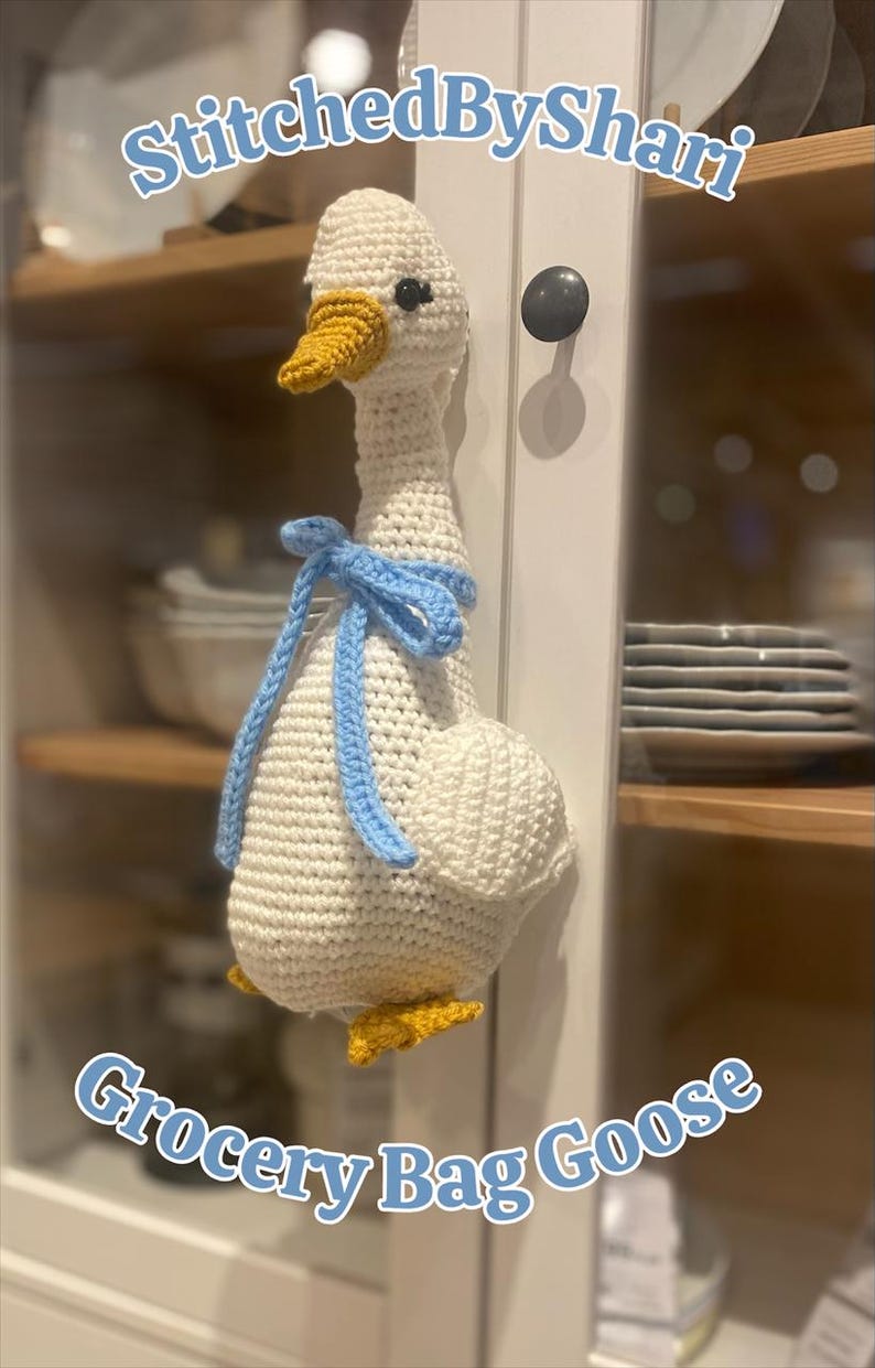 May include: A handmade, crocheted grocery bag goose in white with a yellow beak and feet. It has a light blue bow around its neck. The text "StitchedByShari" and "Grocery Bag Goose" are visible.