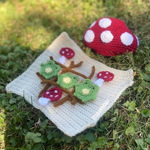 May include: A crocheted tic-tac-toe game with green frog Xs and red mushroom Os on a beige square. A large red mushroom with white spots sits nearby. The game is set on a bed of green grass.