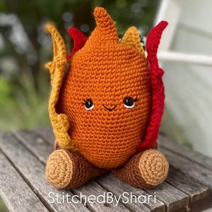 Pode incluir: Um brinquedo de pel&uacute;cia de croch&ecirc; feito &agrave; m&atilde;o em forma de chama. A pel&uacute;cia &eacute; principalmente laranja com detalhes vermelhos e amarelos e apresenta um rosto sorridente com olhos e c&iacute;lios pretos. O texto "StitchedByShari" &eacute; vis&iacute;vel.