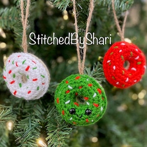 Puede incluir: Tres adornos de donut de ganchillo colgados de un árbol de Navidad. Los donuts son blancos con chispas verdes, rojos con chispas blancas y verdes con chispas blancas. El donut verde tiene una cara sonriente.