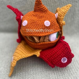 Pode incluir: Um monstro de croch&ecirc; feito &agrave; m&atilde;o com detalhes em laranja, vermelho e amarelo. O monstro tem uma boca grande aberta com dentes, um &uacute;nico olho e bolinhas brancas. O texto "StitchedByShari" &eacute; vis&iacute;vel.