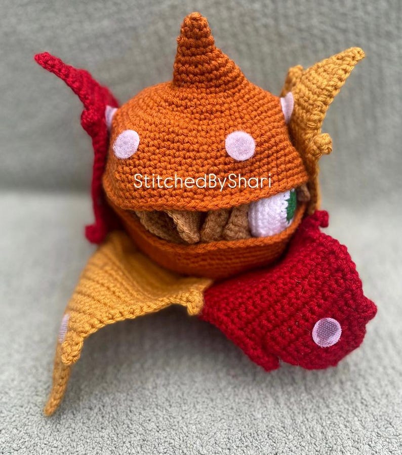 Pode incluir: Um monstro de croch&ecirc; feito &agrave; m&atilde;o com detalhes em laranja, vermelho e amarelo. O monstro tem uma boca grande aberta com dentes, um &uacute;nico olho e bolinhas brancas. O texto "StitchedByShari" &eacute; vis&iacute;vel.
