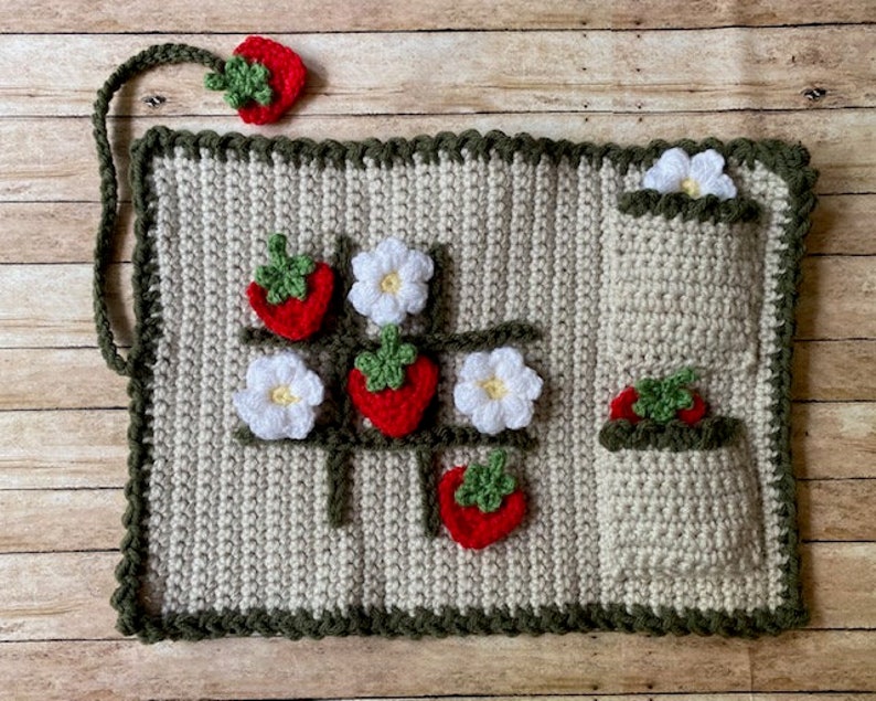 May include: A crocheted strawberry-themed game with a green border. The game features a grid with strawberries and white flowers. There are also pockets on the side for storing the pieces.
