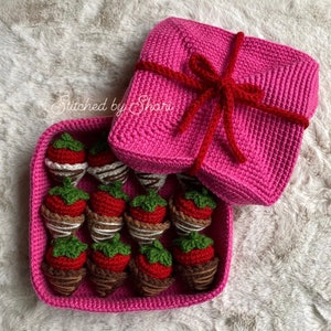 May include: A pink crocheted box with a red ribbon and bow, filled with crocheted chocolate-covered strawberries. The box is open and the strawberries are visible. The text "Stitched by Shari" is visible on the box.