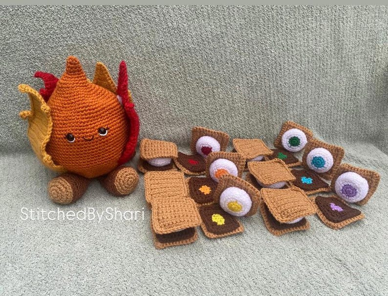 Pode incluir: Uma cole&ccedil;&atilde;o de itens de croch&ecirc; feitos &agrave; m&atilde;o, incluindo uma fogueira laranja sorridente com chamas e v&aacute;rios s'mores. Os s'mores t&ecirc;m v&aacute;rios recheios de marshmallow e chocolate coloridos. O texto "StitchedByShari" &eacute; vis&iacute;vel.