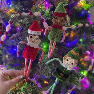 Puede incluir: Tres adornos de elfo de ganchillo hechos a mano con piernas de bastón de caramelo, exhibidos en un árbol de Navidad. Los elfos visten trajes rojos y verdes con sombreros a juego. El texto "Stitched By Shari" es visible en la imagen.
