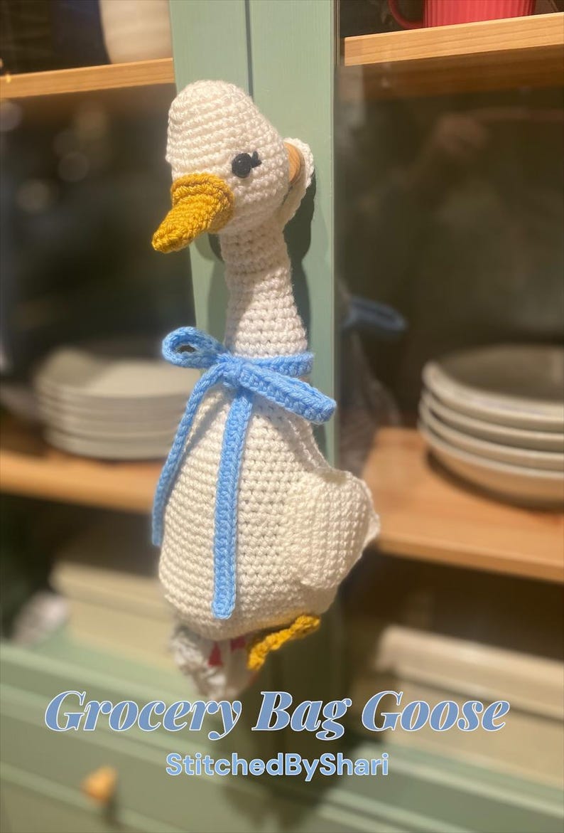May include: Crocheted white goose with a yellow beak and blue bow. The goose is a grocery bag holder. The text "Grocery Bag Goose" and "StitchedByShari" are in the image.