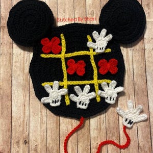May include: A black crocheted Mickey Mouse-shaped tic-tac-toe game. The game board features yellow lines, red bows, and white gloved hands. The text "Stitched By Shari" is visible. A red crocheted string is attached.