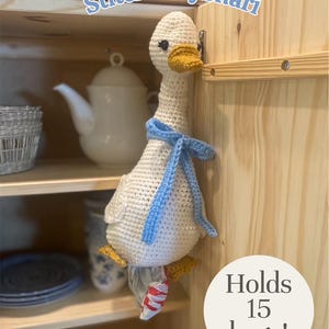 May include: A handmade crochet goose, cream-colored with a yellow beak and feet, and a blue scarf. The goose is holding a plastic bag and is hanging on a wooden cabinet. Text on the image says "Holds 15 bags!" and "StitchedByShari".