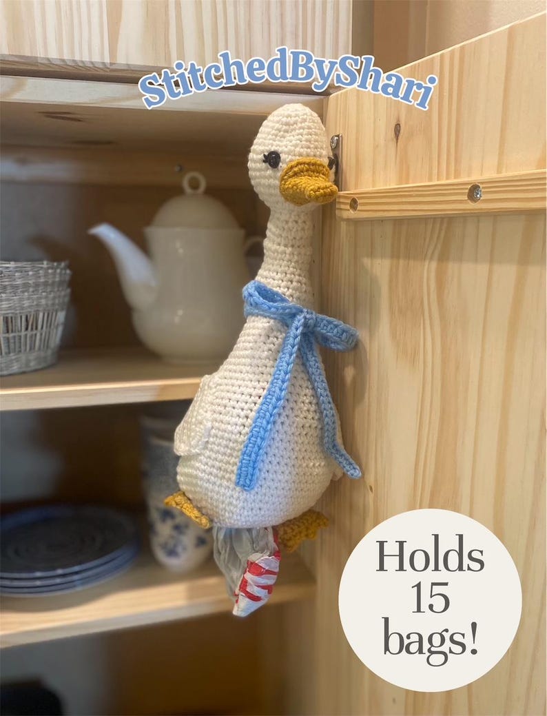 May include: A handmade crochet goose, cream-colored with a yellow beak and feet, and a blue scarf. The goose is holding a plastic bag and is hanging on a wooden cabinet. Text on the image says "Holds 15 bags!" and "StitchedByShari".