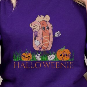 May include: A purple sweatshirt with a cartoon hot dog wearing a chef's hat and holding a mustard bottle. The hot dog is standing in front of a green grass patch with two orange pumpkins. The text 