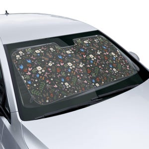 May include: A car windshield sunshade with a dark background and a colorful floral pattern. The pattern includes white daisies, red mushrooms, and blue butterflies. The sunshade is designed to fit the car's windshield.