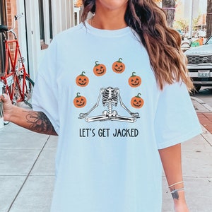 Könnte beinhalten: Weißes T-Shirt mit einem Skelett, das in einer Yoga-Pose sitzt und von Kürbissen umgeben ist. Der Text "Let's Get Jacked" ist auf das Shirt gedruckt.