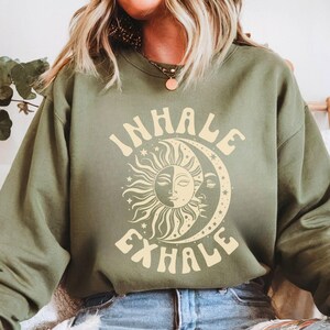 May include: Military green sweatshirt with the words "INHALE EXHALE" in a retro font. The design features a sun and moon graphic. The sweatshirt is a solid color and has long sleeves.