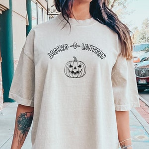以下が含まれることがあります： 薄いベージュのオーバーサイズTシャツに、黒い文字で「Jacked-O-Lantern」と、黒い輪郭でジャック・オ・ランタンが描かれています。Tシャツは柔らかく快適な素材で作られており、アイボリー色があります。