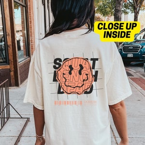 May include: A white t-shirt featuring a graphic of a smiley face formed from the words "Squat, Bench, Dead". The text is in black and orange. The shirt is being worn by a person standing on a pavement.