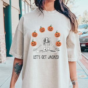 Könnte beinhalten: Ein beigefarbenes Oversize-T-Shirt mit einem Skelett, das Yoga mit Kürbissen um sich herum macht. Der Text auf dem Shirt lautet "Let's Get Jacked".
