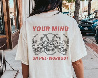 May include: An oversized white t-shirt featuring a graphic of three skulls and the text "YOUR MIND ON PRE-WORKOUT" in red.
