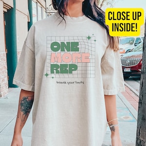 Puede incluir: Una camiseta beige de gran tamaño con un estampado gráfico que dice "ONE MORE REP" en letras verdes y rosas con un patrón de cuadrícula y el texto "break your limits" debajo. La camiseta está puesta en una persona que lleva pantalones cortos negros.