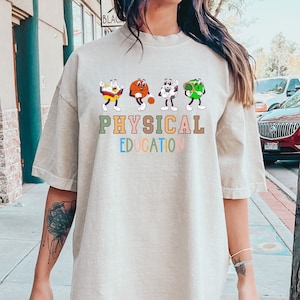 以下が含まれることがあります： 薄いベージュのオーバーサイズTシャツに、スポーツをしている4人の漫画キャラクターのグラフィックがプリントされています。グラフィックの下には「PHYSICAL EDUCATION」という文字がカラフルな文字でプリントされています。シャツは、黒いショートパンツを履いたモデルが着用しています。シャツの色はアイボリーです。