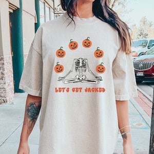 Könnte beinhalten: Ein beigefarbenes Oversize-T-Shirt mit einem Skelett, das mit Kürbissen um sich herum meditiert. Der Text auf dem Shirt lautet "Let's Get Jacked".