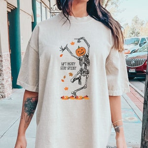 以下が含まれることがあります： かぼちゃの頭をしたスケルトンが踊っているグラフィックがプリントされた、薄いベージュのオーバーサイズTシャツ。シャツのテキストは「Lift Heavy Stay Spooky」です。