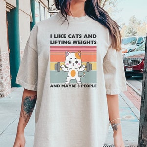 Könnte beinhalten: Ein beigefarbenes Oversize-T-Shirt mit einer Retro-Grafik eines Katzentier, das Gewichte hebt. Der Text auf dem Shirt lautet "Ich mag Katzen und Gewichtheben und vielleicht 3 Leute".