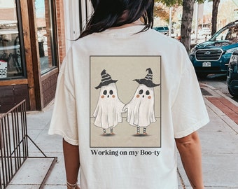 May include: A white t-shirt with a graphic of two ghosts wearing witch hats and the text "Working on my Boo-ty". The shirt is ivory in color.