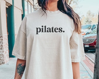 Könnte beinhalten: Ein hellbeiges, übergroßes T-Shirt mit dem Wort "Pilates" in Schwarz auf der Vorderseite. Das Shirt ist aus weichem, bequemem Stoff und hat eine lockere Passform.
