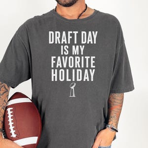 May include: A charcoal gray short-sleeve t-shirt featuring the white text DRAFT DAY IS MY FAVORITE HOLIDAY and a small graphic of a football trophy. The shirt has a crew neckline and a relaxed fit, shown with a brown and white football.