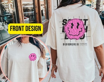 May include: Ivory t-shirt with a pink smiley face graphic on the front and a black and white graphic on the back that says "Squat Bench Dead" with a pink smiley face graphic. The back graphic is on a grid pattern.