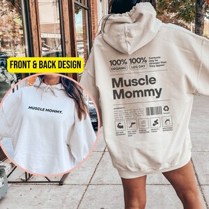 Könnte beinhalten: Ein cremefarbener Hoodie mit dem Text "Muscle Mommy" auf der Vorderseite und "100% Organic 100% Leg Day" und "Muscle Mommy" auf der Rückseite. Der Hoodie hat eine große Tasche auf der Vorderseite und eine Kapuze mit Kordelzug.