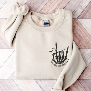 以下が含まれることがあります： 砂色のスウェットシャツに、ロックンロールのハンドジェスチャーをする黒い骨の手が描かれています。手の下には「Lift Heavy Stay Spooky」という文字が印刷されています。