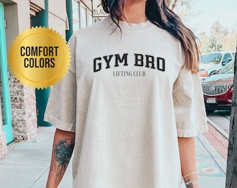 Può includere: Una maglietta oversize color crema con la scritta "GYM BRO LIFTING CLUB" stampata in nero. La modella indossa la maglietta con pantaloncini neri.
