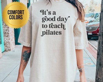 Könnte beinhalten: Eine Frau trägt ein hellbeiges, übergroßes T-Shirt mit dem Aufdruck "It's a good day to teach pilates" in Schwarz. Das Shirt ist locker geschnitten und hat einen Rundhalsausschnitt.