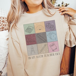 以下が含まれることがあります： ベージュのトレーナーに、9つの正方形のグリッドで構成されたグラフィックデザインが施されています。各正方形には、電卓、分度器、定規、コンパス、鉛筆、そして「MATH」と書かれた本など、数学に関連する異なるオブジェクトが描かれています。グリッドの下には、「IN MY MATH TEACHER ERA」というテキストが印刷されています。