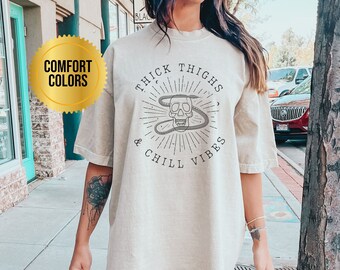 May include: An oversized beige t-shirt featuring the text "Thick Thighs & Chill Vibes" and a skull graphic. The shirt is made from a soft, comfortable material.