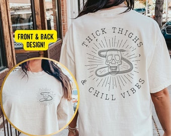 May include: A white t-shirt with a skull and snake design on the back. The text "Thick Thighs & Chill Vibes" is printed on the back of the shirt. The front of the shirt has a smaller version of the skull and snake design.